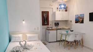 A bedroom with a bed, a desk, a chair, a ceiling fan, and a kitchen area. - Skopelos Cozy Escape Near The Forest the Sea (Skopelos)