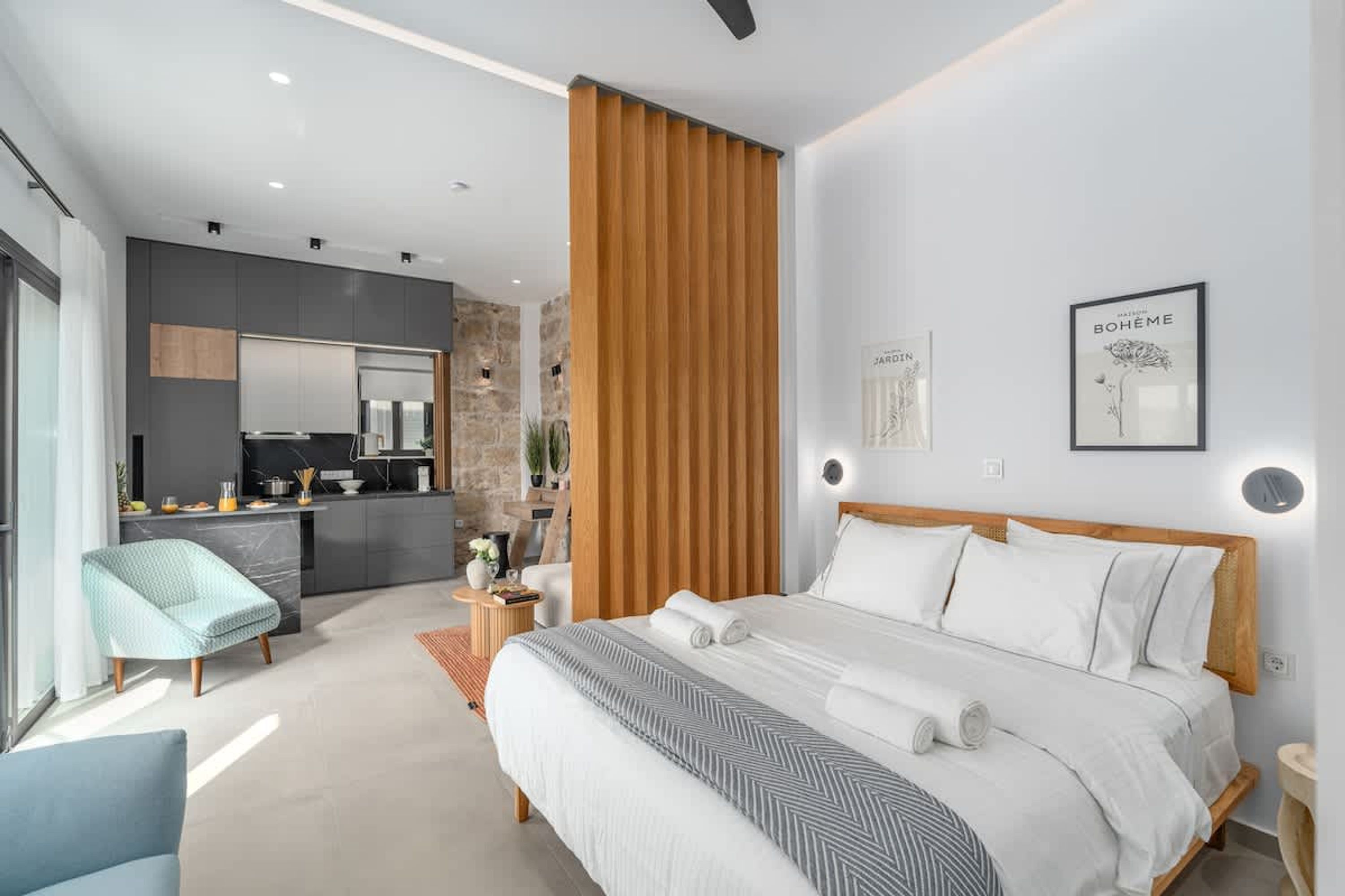 A modern bedroom with a large bed, a wooden partition, a small kitchen area, and a bathroom.