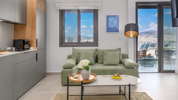 A modern living space with a kitchen, a green sofa, a coffee table, and a view of the city through large windows. - Green Blue Castella Luxury Suites Saronic Pearl (Piraeus)