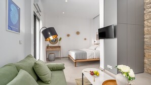 A modern hotel room with a green sofa, a bed with a white headboard, a flat-screen TV mounted on the wall, and a small table with a vase of flowers. - Green Blue Castella Luxury Suites Saronic Pearl (Piraeus)