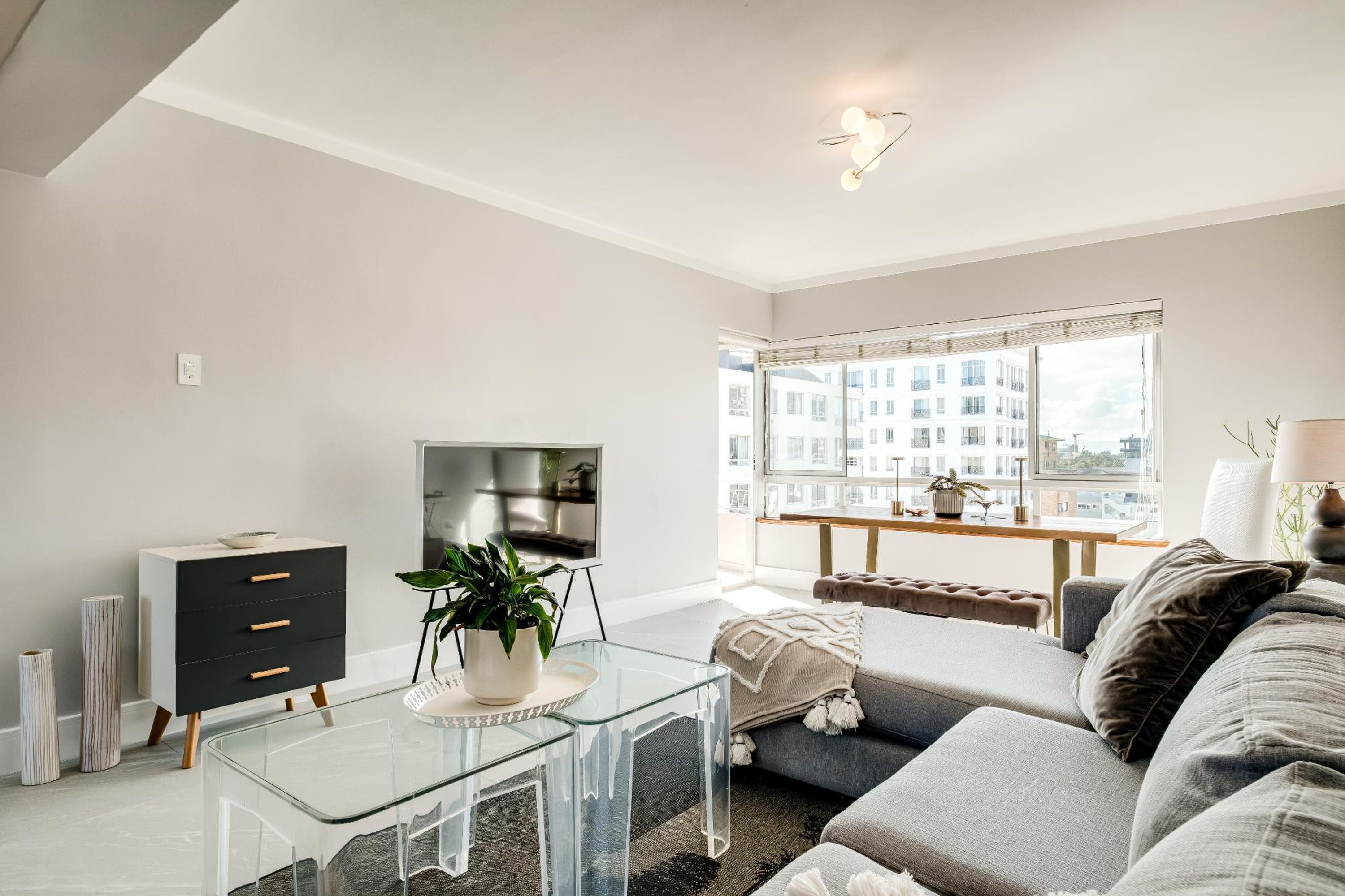 A modern living room with a glass coffee table, a sofa, a TV, and a window with a city view.