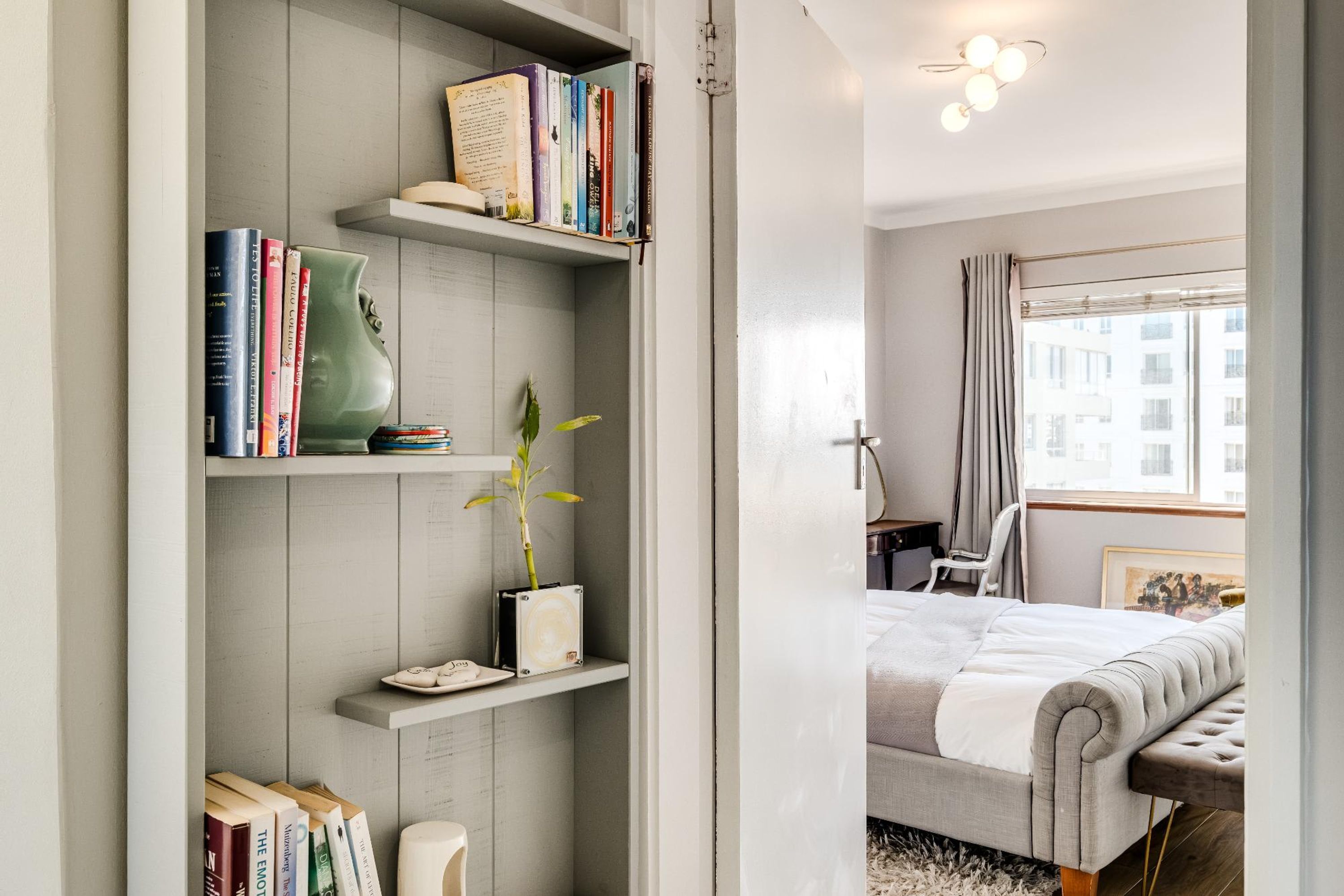 A bedroom with a bed, a bedside table, a chair, a window with curtains, and a bookshelf with books and a plant.