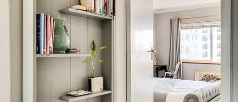 A bedroom with a bed, a bedside table, a chair, a window with curtains, and a bookshelf with books and a plant.