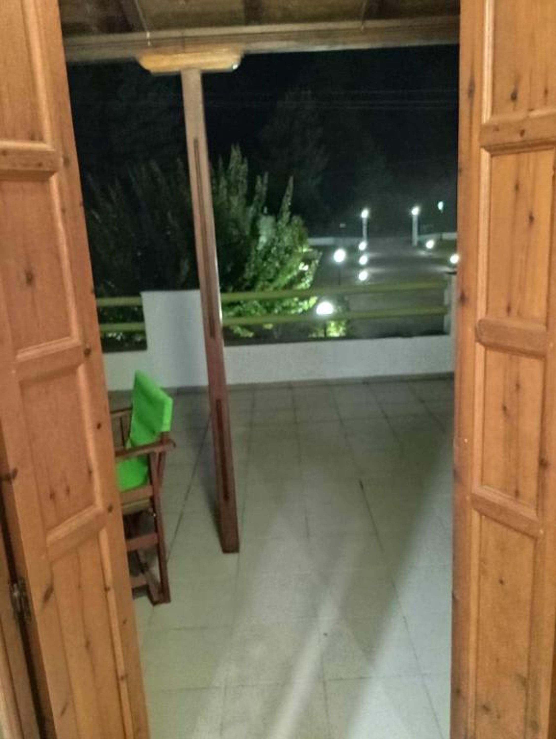 Une porte en bois donne sur un espace extérieur aménagé avec une chaise et une petite table, un sol carrelé et une vue sur une rue illuminée la nuit.