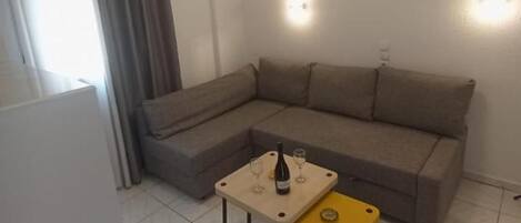 A living room with a sectional sofa, a small table with a bottle and glasses, and a yellow ottoman.