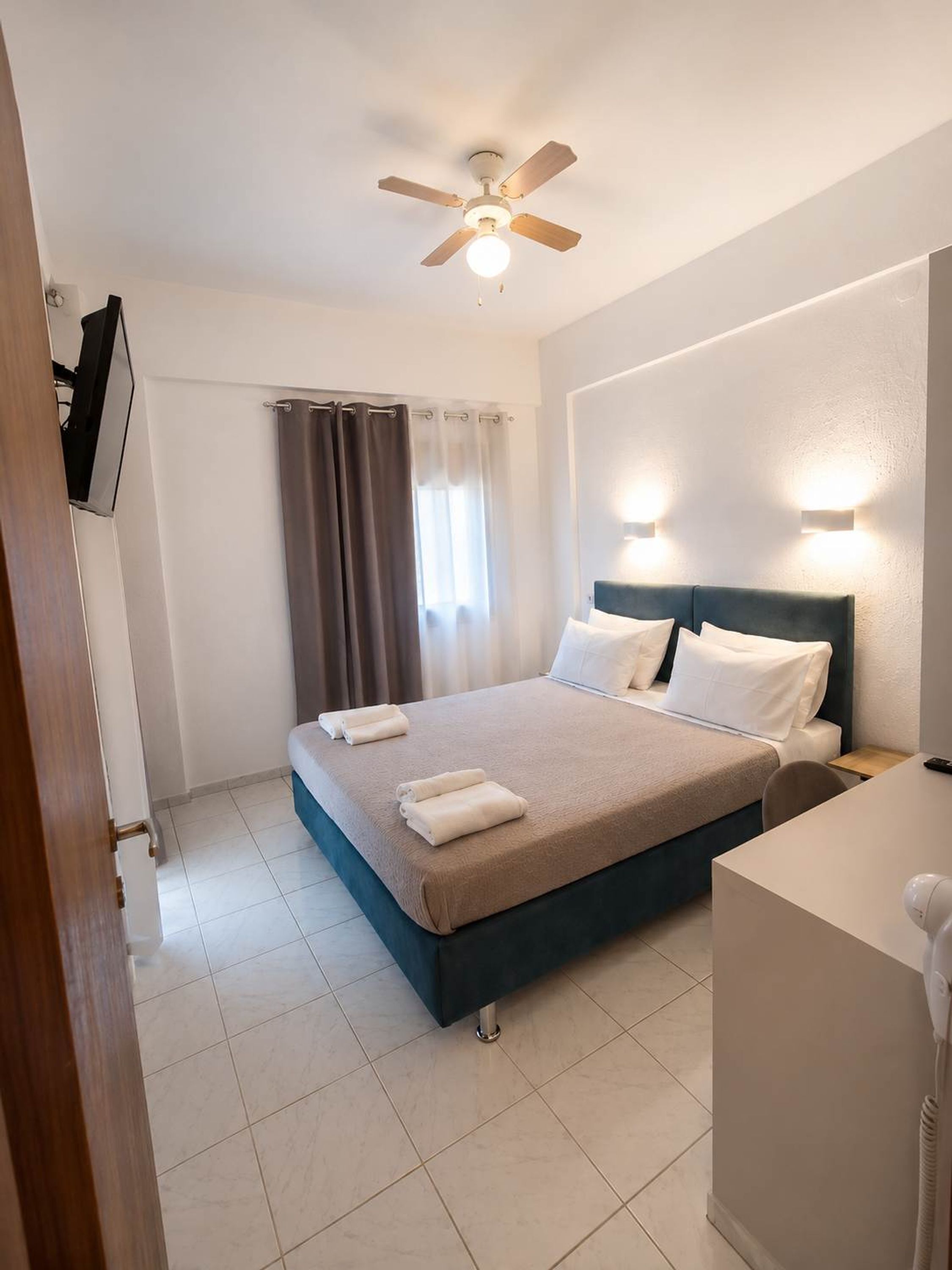 A hotel room with a bed, a desk, a chair, a ceiling fan, a window with curtains, and a wall-mounted light fixture.