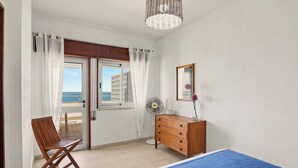 Quarto com cama, cómoda de madeira, cadeira, ventoinha e vista para o mar através de uma janela.