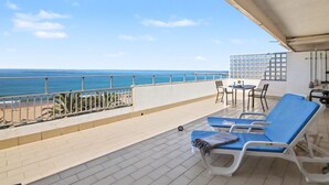 Uma varanda com vista para o mar, uma mesa com cadeiras e dois espreguiçadeiras.
