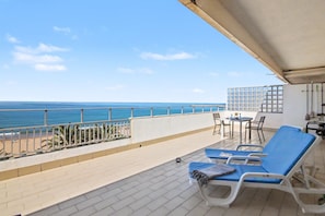 A balcony with a view of the sea, a table with chairs, and two sun loungers.