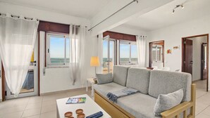 Uma sala de estar com sofá, mesa de centro e vista para o mar através de grandes janelas.