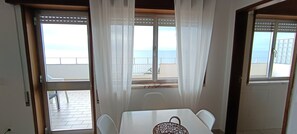 Une piĂšce avec une table et des chaises, une vue sur la mer par une fenĂȘtre et un balcon avec une balustrade.