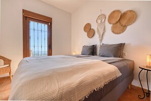 Een slaapkamer met een bed, een nachtkastje, een macramé wanddecoratie en uitzicht door een raam met een houten deur.
