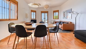 A dining area with a round table, chairs, and a view of the sea through large windows. - Kousgaard - Spacious private villa in quiet area with pool and sea view (Sayalonga)