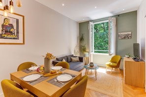 Ein Essbereich mit Tisch für zwei Personen, einer Couch, einem Fernseher und einem Fenster mit Blick auf Grünflächen.