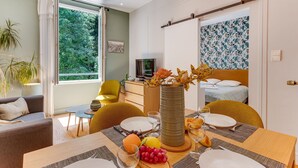 Ett rum med ett matbord för två personer, en säng med sänggavel, en tv och ett stort fönster med gardiner.