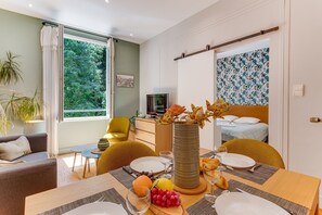 Ein Zimmer mit Esstisch für zwei Personen, einem Bett mit Kopfteil, einem Fernseher und einem großen Fenster mit Vorhängen.