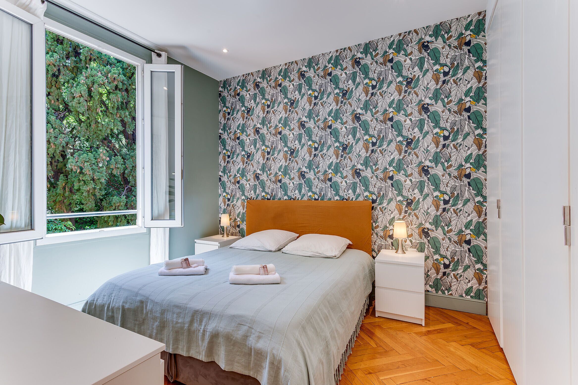 A bedroom with a large window, a bed with a wooden headboard, two bedside tables with lamps, and a patterned wall.