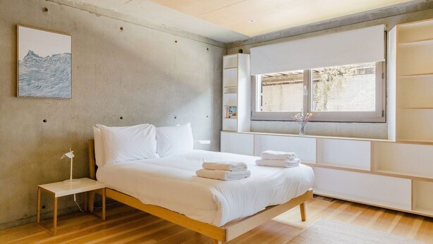 Quarto moderno com cama de madeira, mesa de cabeceira, janela com vista e prateleira na parede.