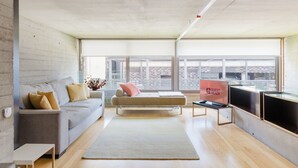 Uma sala de estar moderna com um sofá, uma mesa de centro e uma TV. Há uma janela grande com vista para edifícios.
