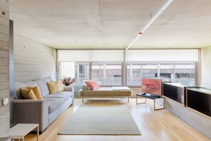 Ein modernes Wohnzimmer mit einer Couch, einem Couchtisch und einem Fernseher. Ein großes Fenster bietet Blick auf Gebäude.