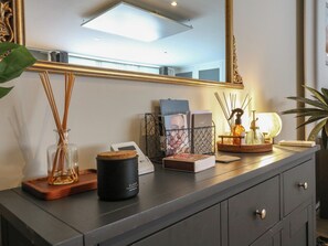 A dark-colored dresser with a mirror, a candle, a diffuser, and a basket with a photo frame and books. - Denmark House (Chatham)