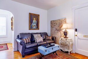 Ein Wohnzimmer mit einem dunkelbraunen Ledersofa, gemusterten Kissen, einem blauen Hocker, einem kleinen weißen Beistelltisch mit einer Lampe und einem großen gerahmten Gemälde an der Wand.