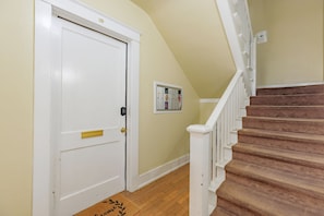 A hallway with a white door, a doormat, a staircase with a white railing, and a wall-mounted bulletin board.