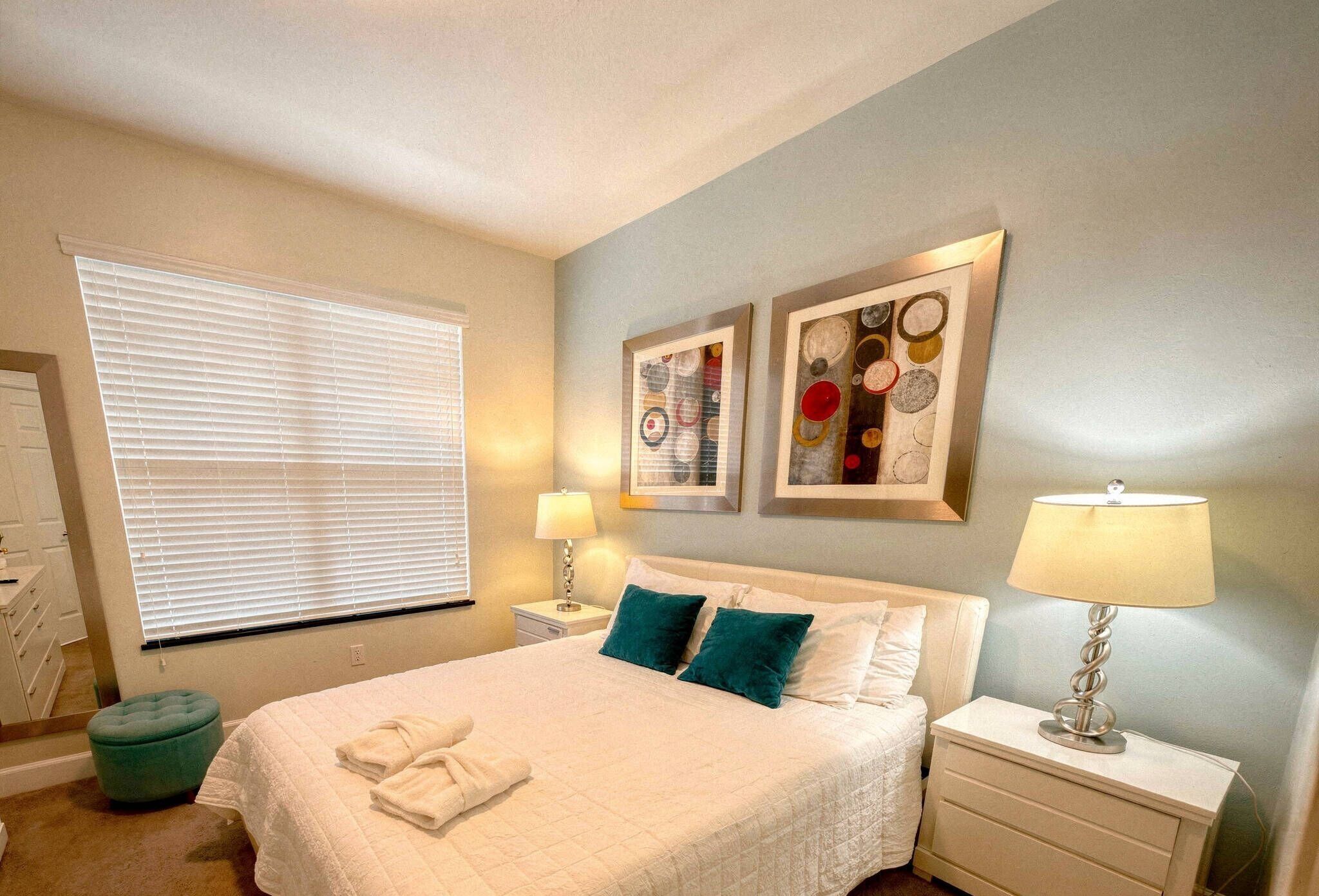 A bedroom with a bed, two nightstands, a green ottoman, and artwork on the wall.