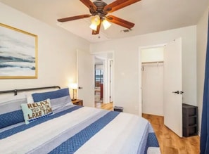 Un dormitorio con cama, ventilador de techo, puerta que da a otra habitación y un cuadro en la pared.
