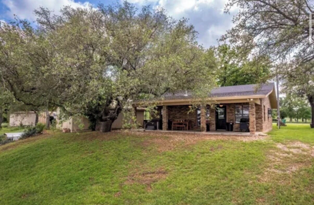 A stone building with a covered patio, surrounded by trees and a grassy lawn.