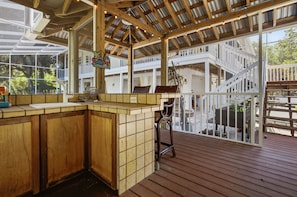 Ein überdachter Außenbereich mit Holzdeck, einer Bar mit Theke und Hockern sowie Blick auf ein weißes Gebäude mit Balkonen.