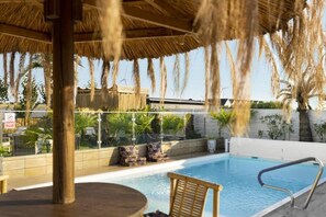 A pool area with a thatched roof, lounge chairs, and a view of palm trees. - Holiday home with terrace, garden, and pool (Kopan)