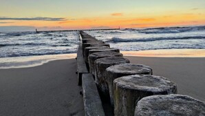 A beach with a wooden jetty extending into the ocean, waves crashing against the shore, and a sunset in the background. - Holiday home with garden, air conditioning, pool (Kopan)