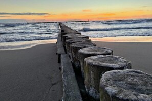 Ein Strand mit einer hölzernen Mole, die sich ins Meer erstreckt, Wellen schlagen an die Küste, und ein Sonnenuntergang im Hintergrund.
