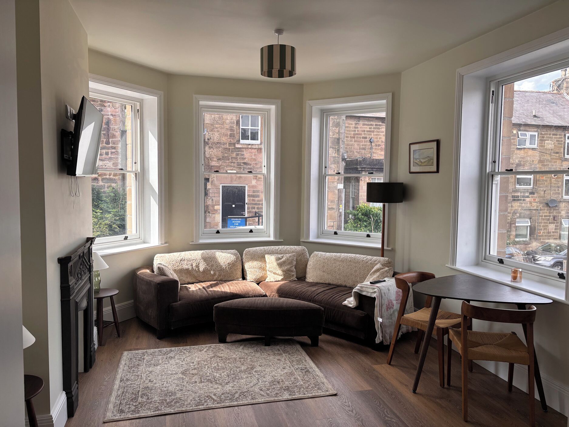 A cozy living room with a sofa, ottoman, and a dining table. There are three windows providing views of the outside.
