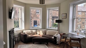 A cozy living room with a sofa, ottoman, and a dining table. There are three windows providing views of the outside. - Holt Hall - Ground Floor Apartment (Matlock)