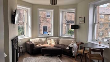 A cozy living room with a sofa, ottoman, and a dining table. There are three windows providing views of the outside.