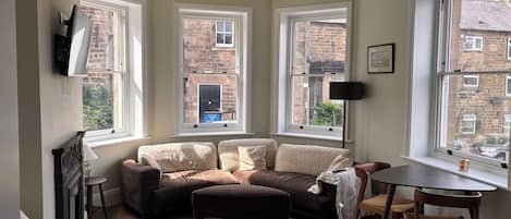 A cozy living room with a sofa, ottoman, and a dining table. There are three windows providing views of the outside.