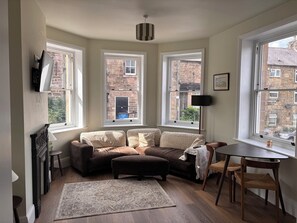 Ein gemütliches Wohnzimmer mit Sofa, Hocker und Esstisch. Drei Fenster bieten Ausblicke nach draußen.