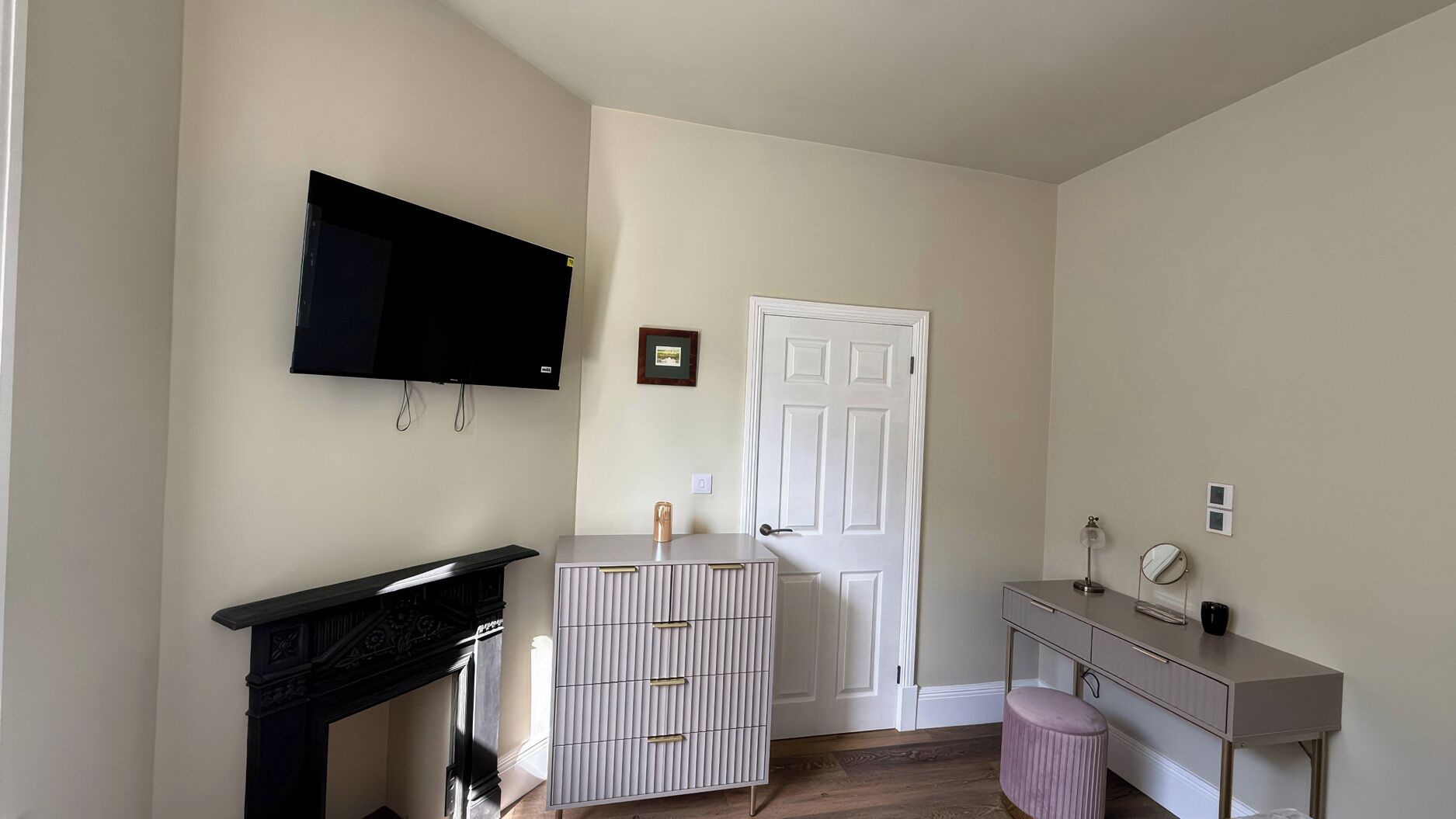 Ein Zimmer mit flachbildfernseher an der Wand, einer weißen Tür, einer kleinen Kommode und einem Schminktisch mit Spiegel sowie einem pinken Hocker.