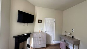 A room with a flat-screen TV mounted on the wall, a white door, a small cabinet with drawers, a vanity with a mirror, and a pink stool. - Holt Hall - Ground Floor Apartment (Matlock)
