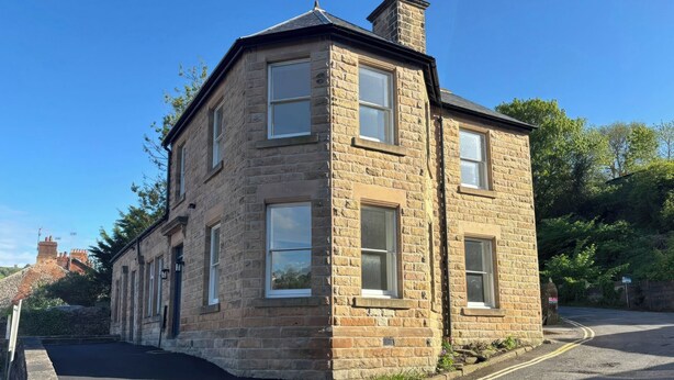 A stone building with a corner entrance, a paved driveway, and a clear sky. - Holt Hall - Ground Floor Apartment (Matlock)