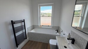 Ein Badezimmer mit einer Badewanne, einem Waschbecken, einer Toilette, einem Handtuchhalter und einem Fenster mit Blick auf eine Stadtlandschaft.