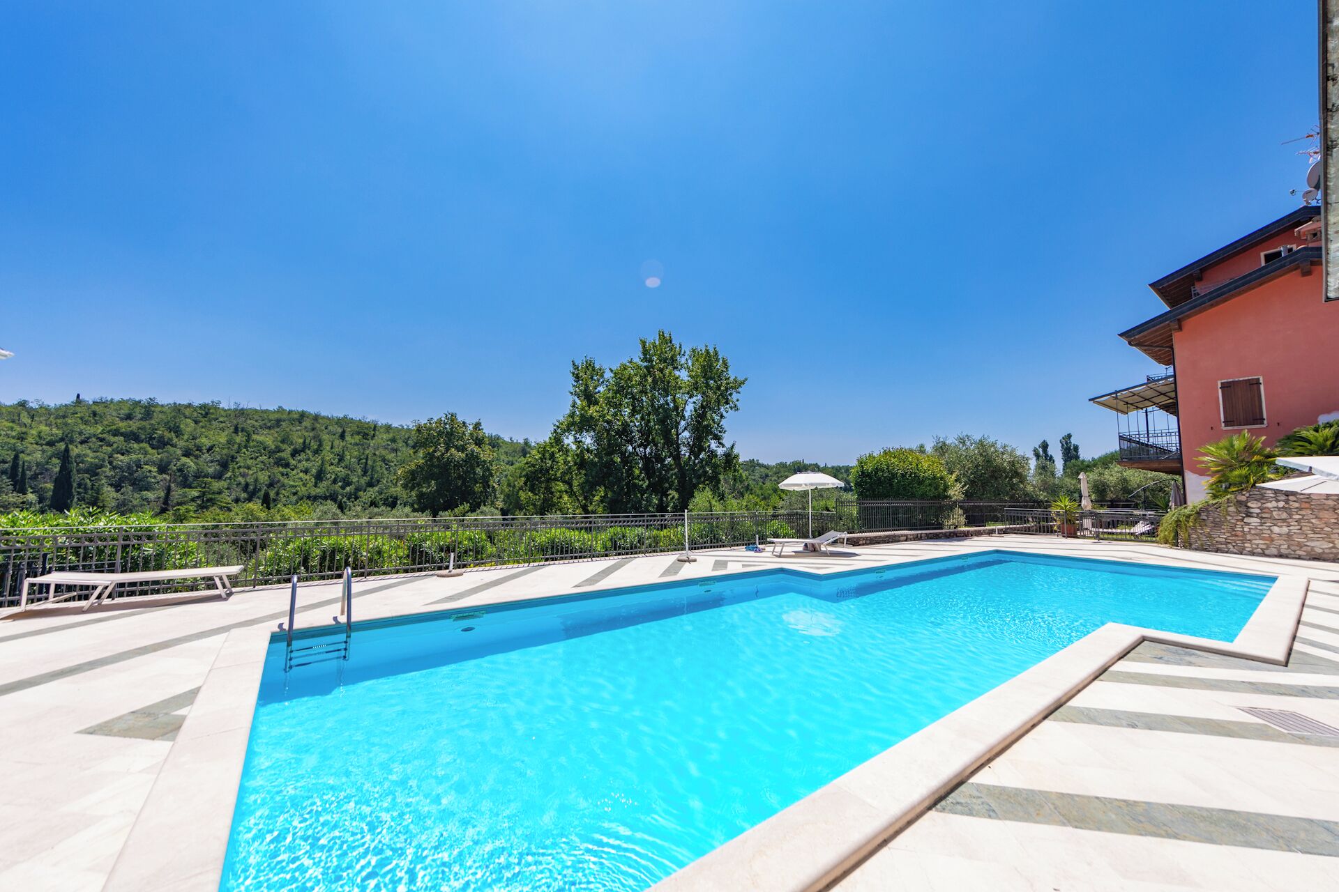 Una piscina rettangolare con acqua cristallina e blu, circondata da un solarium piastrellato, una recinzione in metallo e un edificio rosso sullo sfondo.