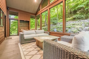 Ein modernes Interieur mit Rattanmöbeln, großen Fenstern und Blick auf eine bewaldete Landschaft.