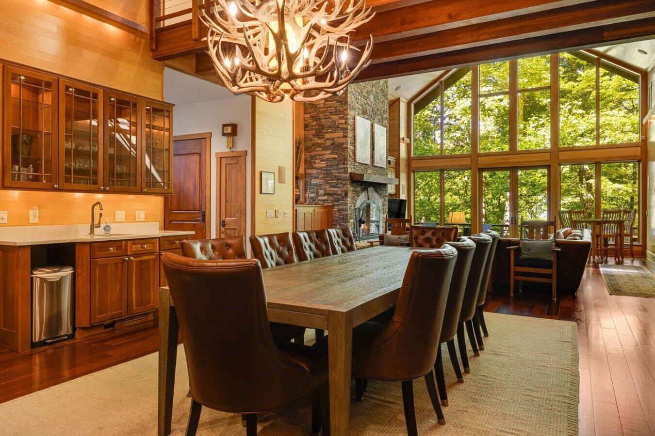 A spacious dining area with a large wooden table and leather chairs, a stone fireplace, and large windows offering a view of the outdoors.