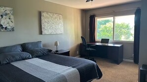 Un dormitorio con una cama, un escritorio con una computadora portátil, una silla, un ventilador de techo y obras de arte en las paredes.