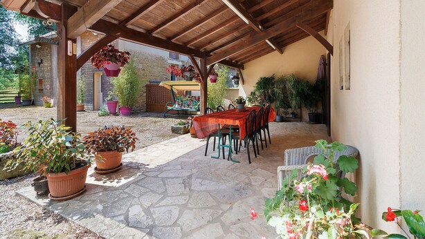 Une terrasse couverte avec un sol en pierre, un plafond en bois et une table entourée de chaises.