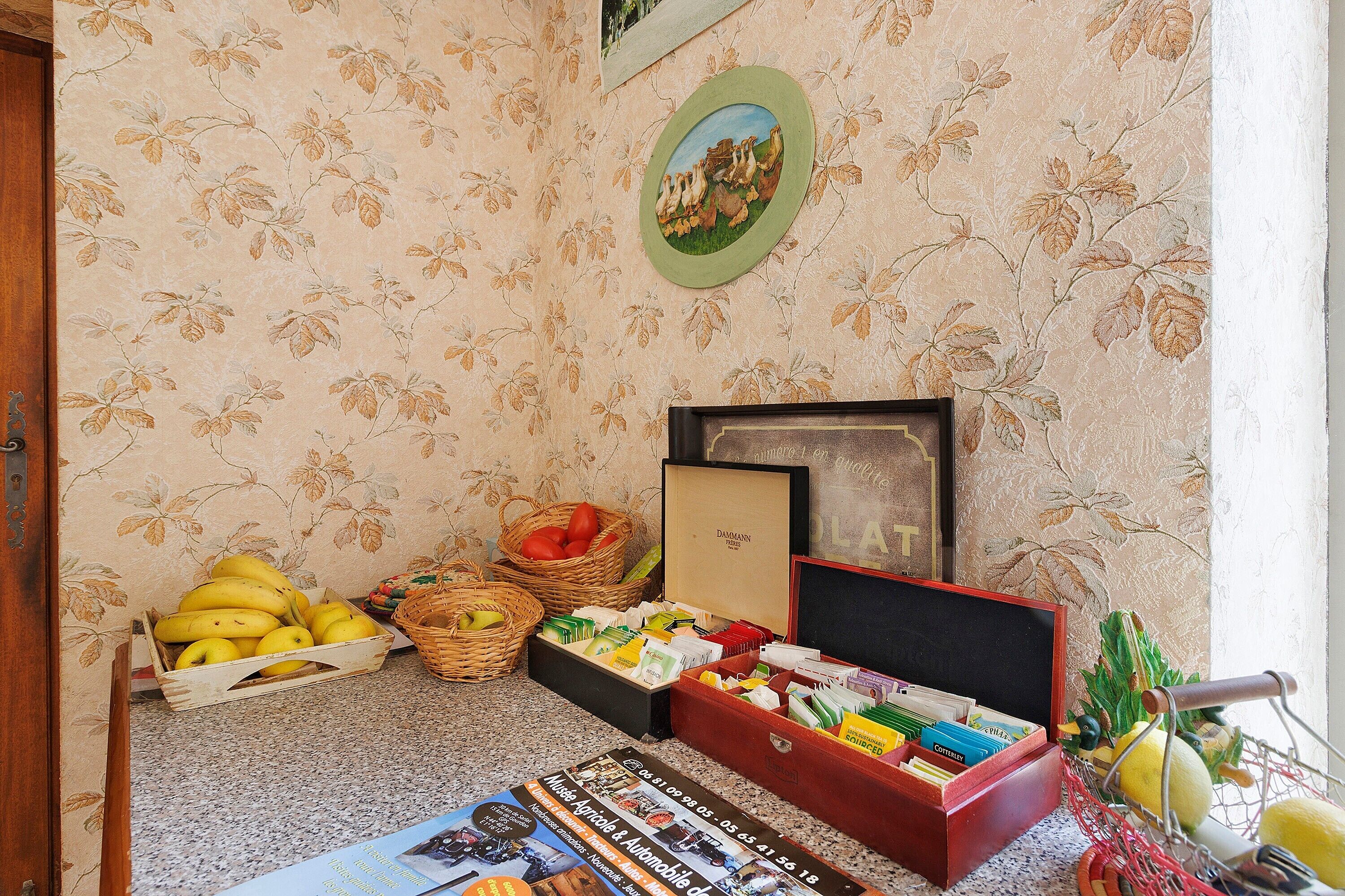 A room with floral wallpaper, a basket of bananas, a wicker basket with tomatoes, a red box with books, and a framed picture on the wall.
