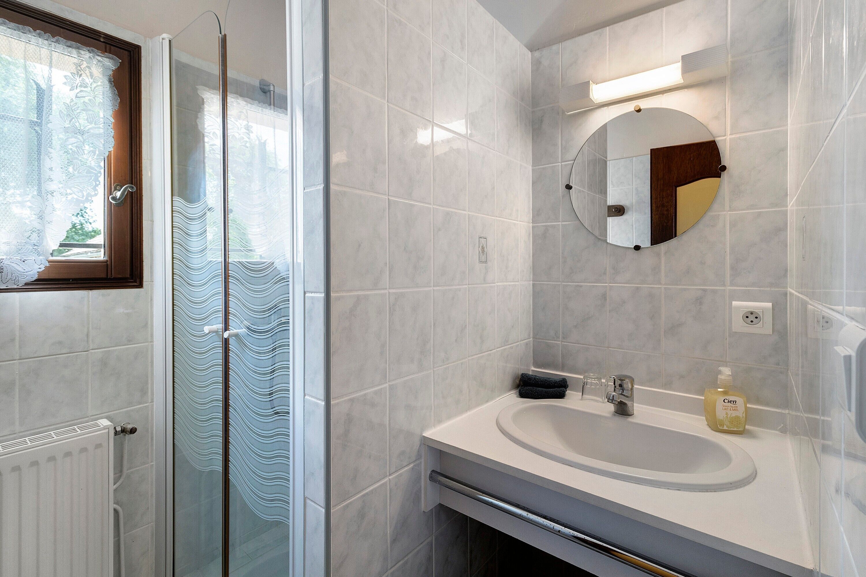Ein Badezimmer mit Duschkabine aus Glas, Waschbecken mit Spiegel und Fenster mit Spitzenvorhängen.
